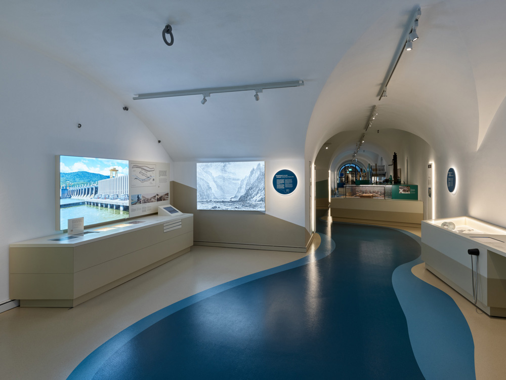 Blick durch Museumsräume. Der Boden visualisiert die Gestaltungsidee und leitet durch die Ausstellung: Geschwungene Intarsie, in der Mitte blau, am Rand beige, symbolisiert den Flusslauf. Seitlich Schau-Leuchttafeln